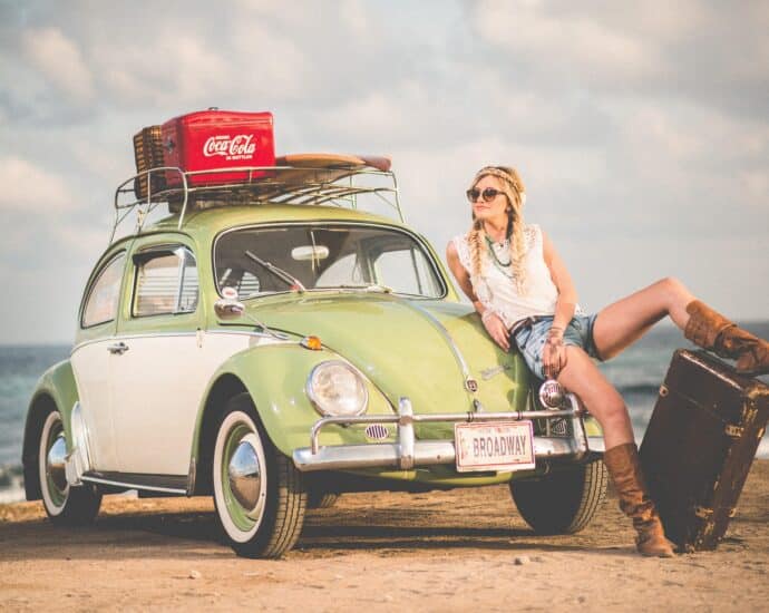 A blonde woman leaning against the bonnet of an old style light green Beetle on a beach. The registration plate has Broadway written on it and there's a case of Coca Cola on the roof of the car