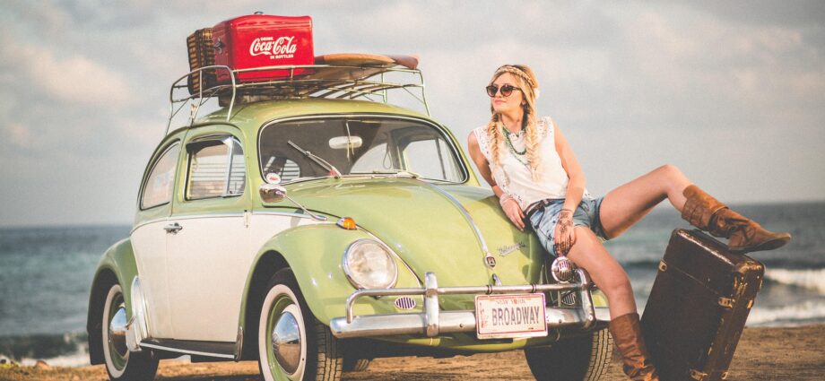 A blonde woman leaning against the bonnet of an old style light green Beetle on a beach. The registration plate has Broadway written on it and there's a case of Coca Cola on the roof of the car